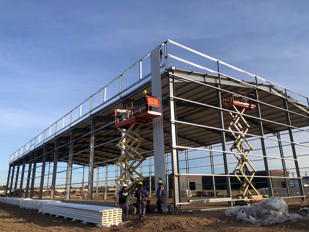 Estructura metálica de una nave industrial en montaje, con dos plataformas elevadoras y operarios trabajando